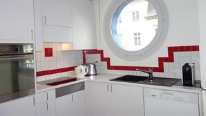Uma cozinha moderna com armários brancos, painel de azulejos vermelhos, forno embutido, pia e janela com vista para edifícios.