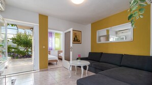 Ein Wohnzimmer mit grauem Sofa, einem kleinen weißen Tisch und einem Bett mit weißer Bettwäsche. An der Wand hängt ein Gemälde und über dem Sofa eine Pflanze.