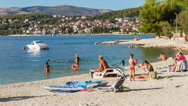 A beach with people sunbathing and swimming, a boat on the water, and a town in the background. - DOROTEA 1, 3-room apartment for 6 persons, 50m from the pebble beach (Okrug Gornji)
