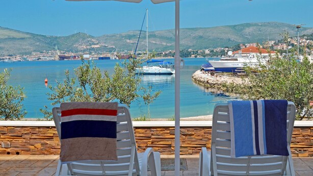 Deux fauteuils de jardin blancs, accompagnés de serviettes, offrent une vue sur une marina où sont amarrés des bateaux, ainsi que sur une ville côtière.