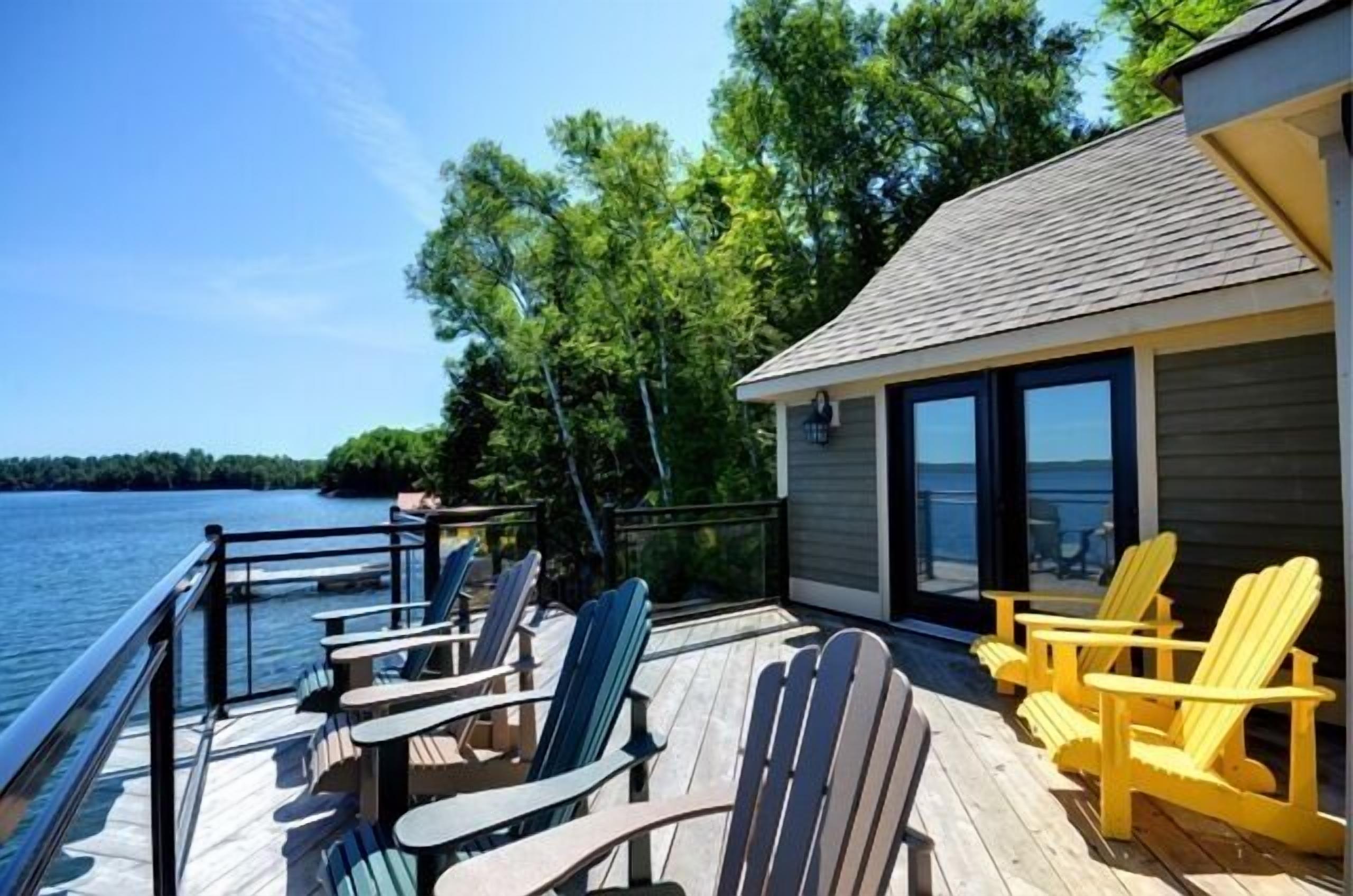 A deck with Adirondack chairs and tables overlooking a lake, with a building adjacent to the deck.