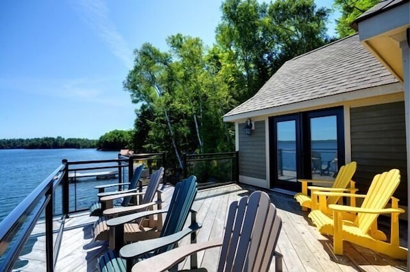 A deck with Adirondack chairs and tables overlooking a lake, with a building adjacent to the deck.