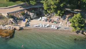 Luftaufnahme eines Strandes mit klarem Wasser, weißen Liegestühlen und einem Steinweg, der zum Wasser führt.
