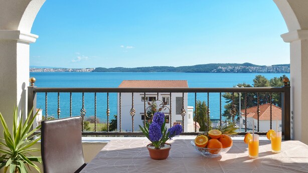 Ein Balkon mit Meerblick, ein Tisch mit Früchten und einem Getränk sowie eine Topfpflanze.