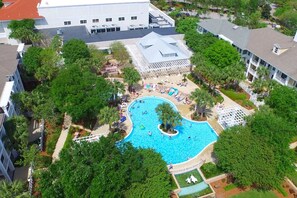 An aerial view of a resort with a central swimming pool surrounded by lush greenery, a white building with multiple windows, and a covered area with umbrellas. - Charming Baytowne Wharf 1BR/2BA + Bunk Room — Steps to Village (Destin)