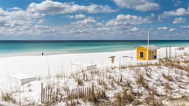 A beach scene with a small yellow building, white sand, and a clear blue sky.