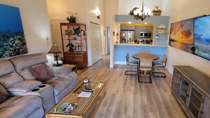 A living room with a sofa, coffee table, and TV. There is a dining area with a table and chairs. A kitchen is visible in the background.
