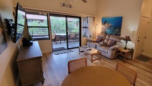 A living room with a sofa, coffee table, and chairs, a television, a painting, and a view of a balcony with plants.
