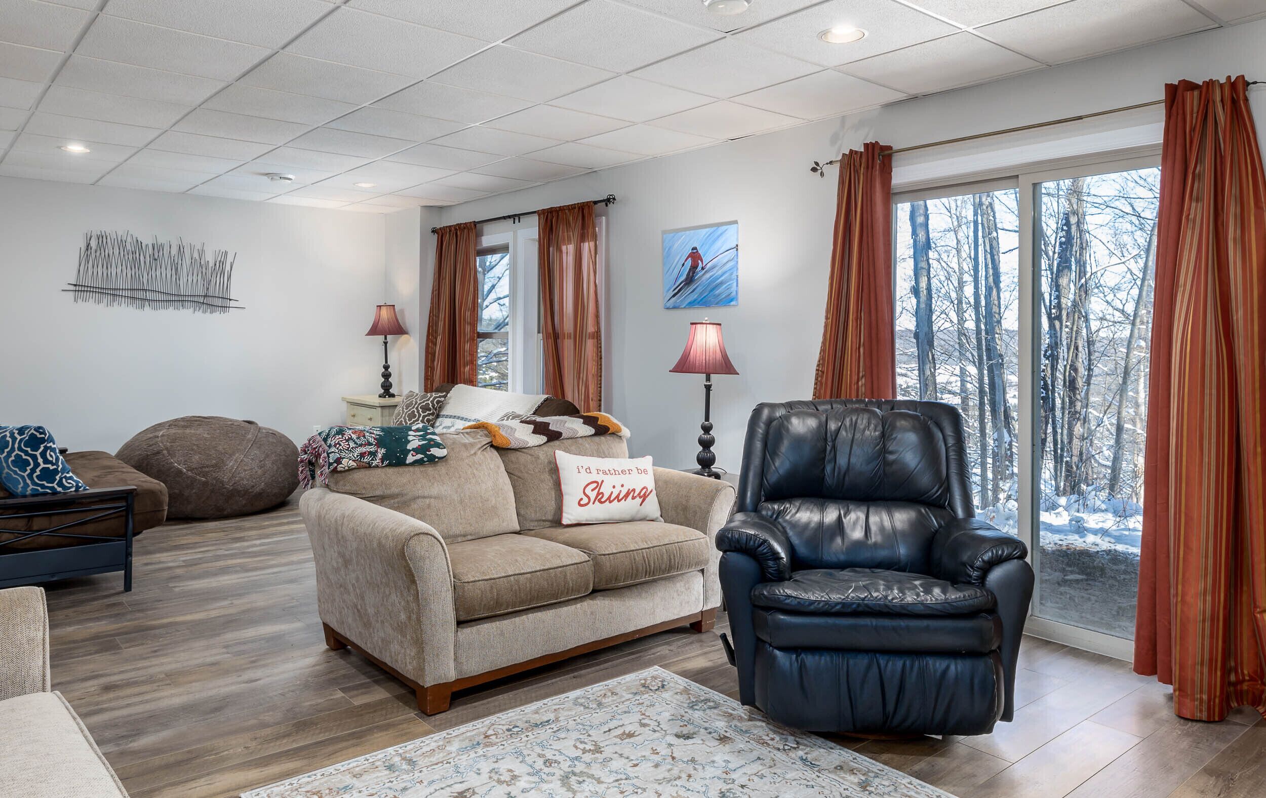 A cozy living room with a beige sofa, a black leather chair, and a large window with curtains.