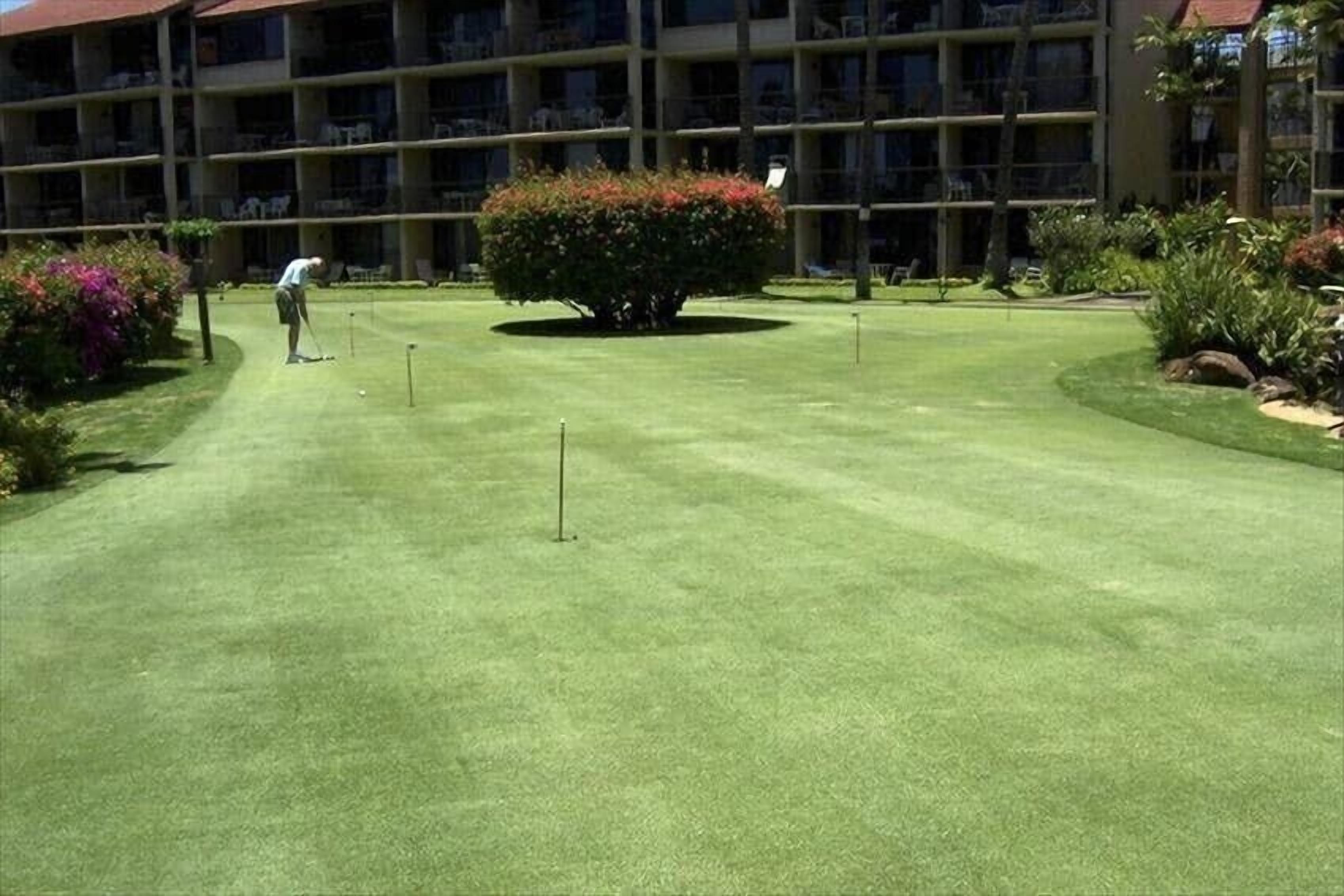 A well-maintained golf course with a putting green, surrounded by lush landscaping and a multi-story building in the background.