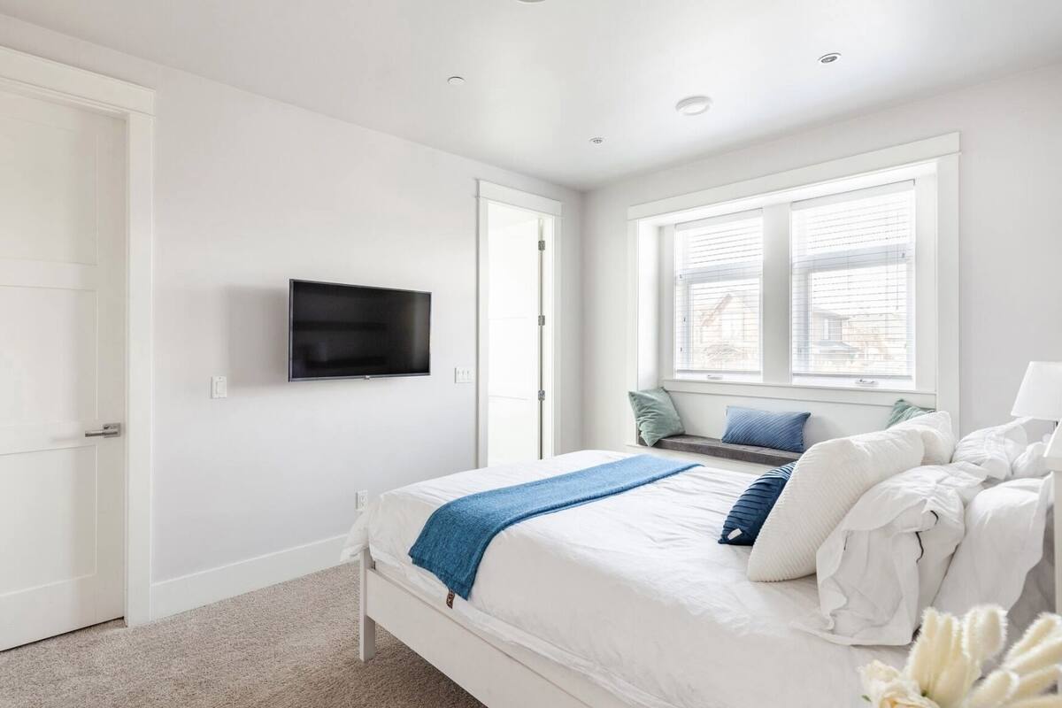 A bedroom with a bed, a TV mounted on the wall, a window with blinds, and a door.