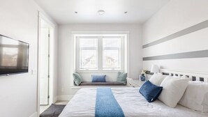 Een slaapkamer met een bed, een raam met zonwering, een televisie aan de muur en een nachtkastje met een lamp en een vaas.