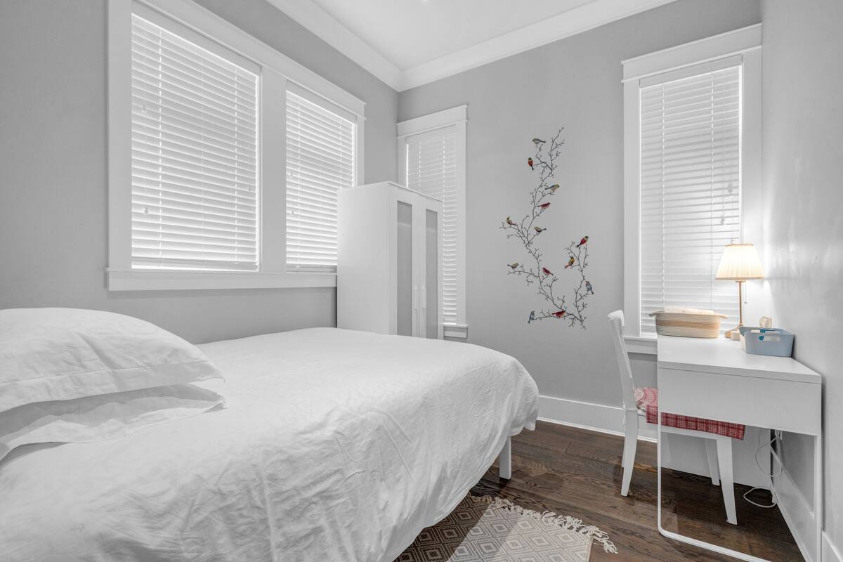A bedroom with a bed, a desk, and a chair. There are windows with blinds, a wall decoration, and a lamp on the desk.