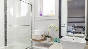 Een moderne badkamer met een douchecabine van glas, een wit toilet, een wastafel met spiegel erboven en een raam dat voor natuurlijk licht zorgt.