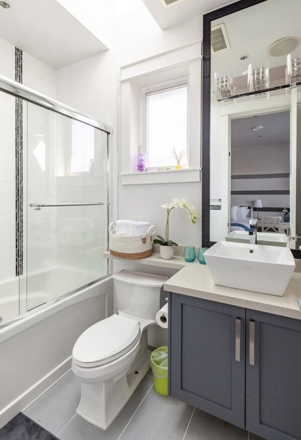 A modern bathroom with a glass-enclosed shower, a white toilet, a sink with a mirror above, and a window providing natural light. - Vancouver West Cozy Retreat by Host Launch (Vancouver)