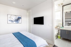 A modern bedroom with a bed, a flat-screen TV, a painting, and a bathroom with a sink and mirror. - Vancouver West Cozy Retreat by Host Launch (Vancouver)
