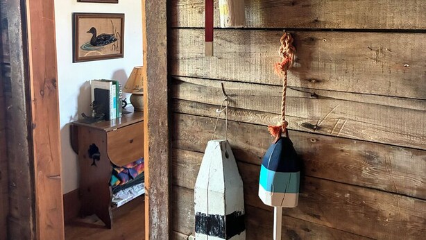 A wooden cabin interior with a door leading to a room, a framed painting of a boat, and a small table with a lamp and books.