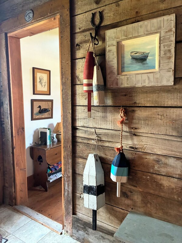 L’intérieur d’un chalet en bois, avec une porte donnant sur une pièce, un tableau encadré représentant un bateau, et une petite table sur laquelle se trouvent une lampe et des livres.