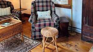 Une pièce chaleureuse avec un plafond en bois, un fauteuil à carreaux, une petite table d’appoint en osier, un petit tabouret et une armoire en bois.