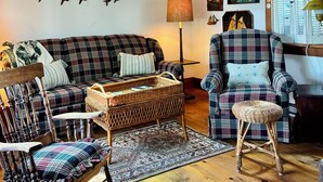 A cozy living room with a wooden ceiling, a rocking chair, a plaid sofa, and a chair with a woven seat.
