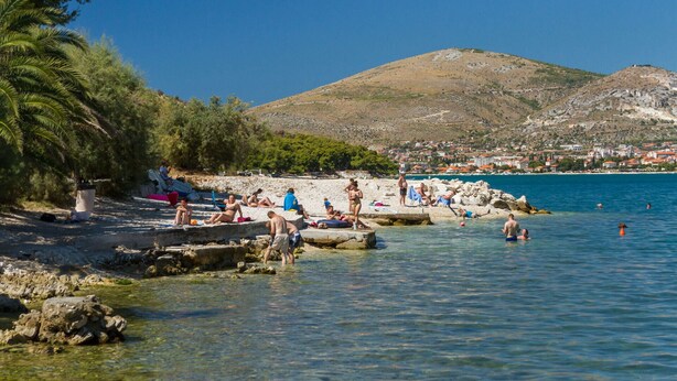 Ein Strand mit klarem Wasser, badenden Personen und einem Berg im Hintergrund.