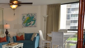 A living room with a blue sofa, white chairs, a coffee table, a ceiling fan, and a large window with a view of buildings. - Kick back, relax, and catch a wave! (Destin)