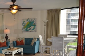 A living room with a blue sofa, white chairs, a coffee table, a ceiling fan, and a large window with a view of buildings. - Kick back, relax, and catch a wave! (Destin)