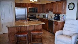 A kitchen with wooden cabinets, a stainless steel refrigerator, a microwave, a stove, and a coffee maker. There are two bar stools and a chair. - Kick back, relax, and catch a wave! (Destin)