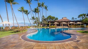 A pool area with lounge chairs and umbrellas, surrounded by palm trees and a building with a roofed patio. - Wailea Ekahi 45B-Newly Remodeled Kitch, Large Lanai, Pool #3 w/BBQ (Kihei)