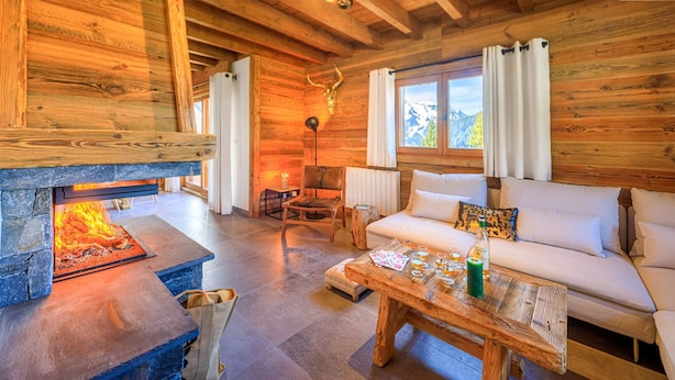 A cozy log cabin interior with a stone fireplace, a sofa, a coffee table, and a view of mountains through the windows. - Modern chalet 10pers - close to slopes and center (Châtel)