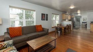 A spacious living area with a sofa, coffee table, and dining table. There are large windows providing natural light and a view of the outdoors. - West Side Gem │King Beds │Walk Everywhere │Hot Tub │Large Living Spaces (Bend)