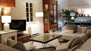 A spacious living room with a high ceiling, a television, a glass coffee table, a cabinet, and a sofa. - Former sheepfold with swimming pool, among the pines, near the sea. (Aude)