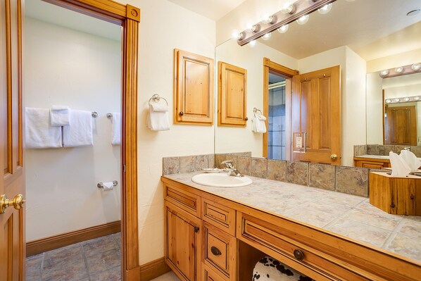Une salle de bain avec un meuble-lavabo en bois, un évier et un miroir avec éclairage. Des serviettes sont accrochées au mur et une porte donne accès à une autre pièce.