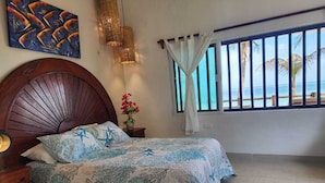 Un dormitorio con cama, cabecera de madera, mesita de noche, un florero con flores, un cuadro en la pared y ventana con vista al océano.