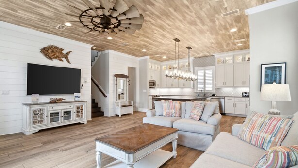 A spacious living room with a large flat-screen TV, a wooden coffee table, and a sofa with colorful pillows. The room features a wooden ceiling and white walls.