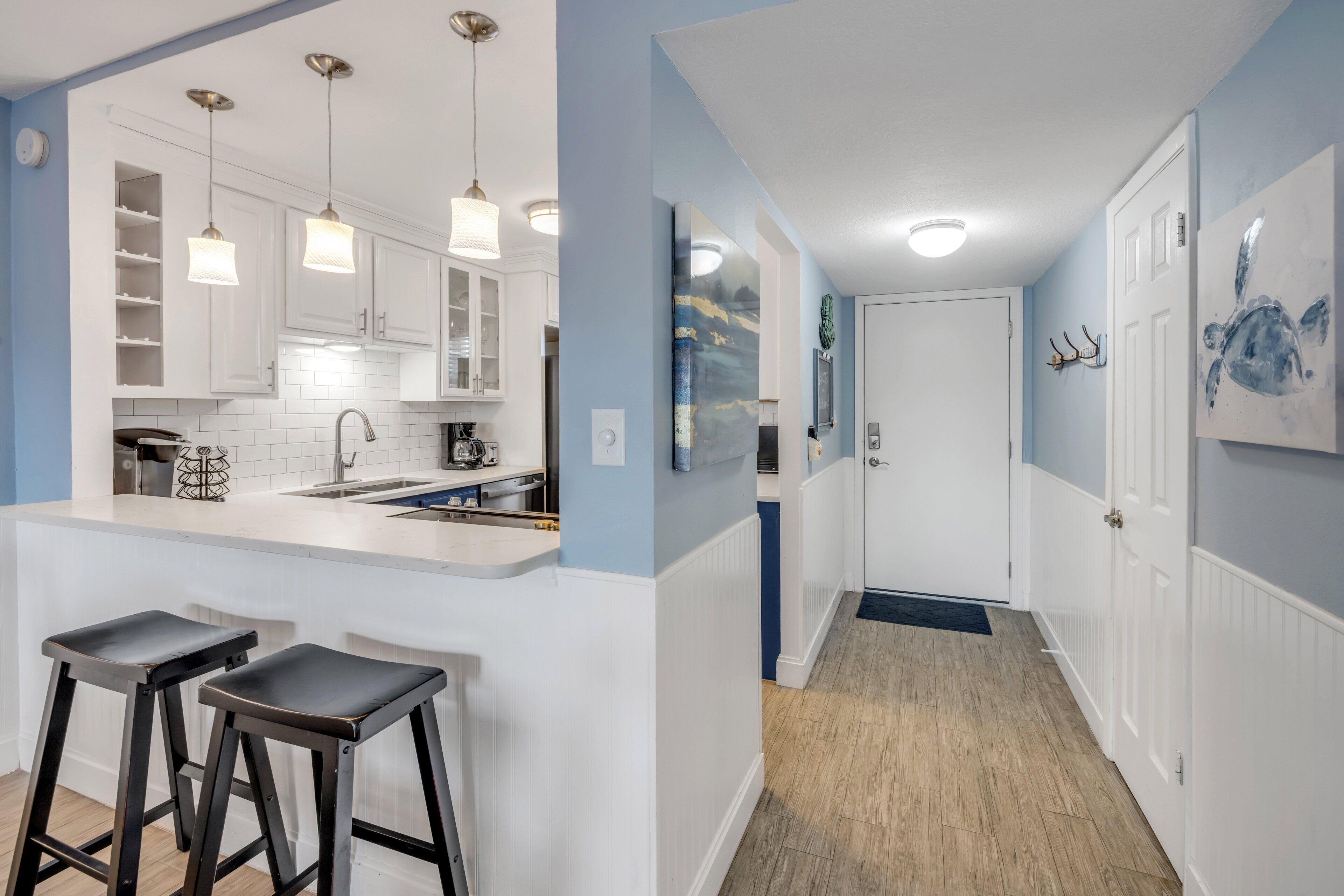 Una cocina moderna con armarios blancos, una isla central con taburetes, iluminación colgante y un pasillo con una puerta y obras de arte.