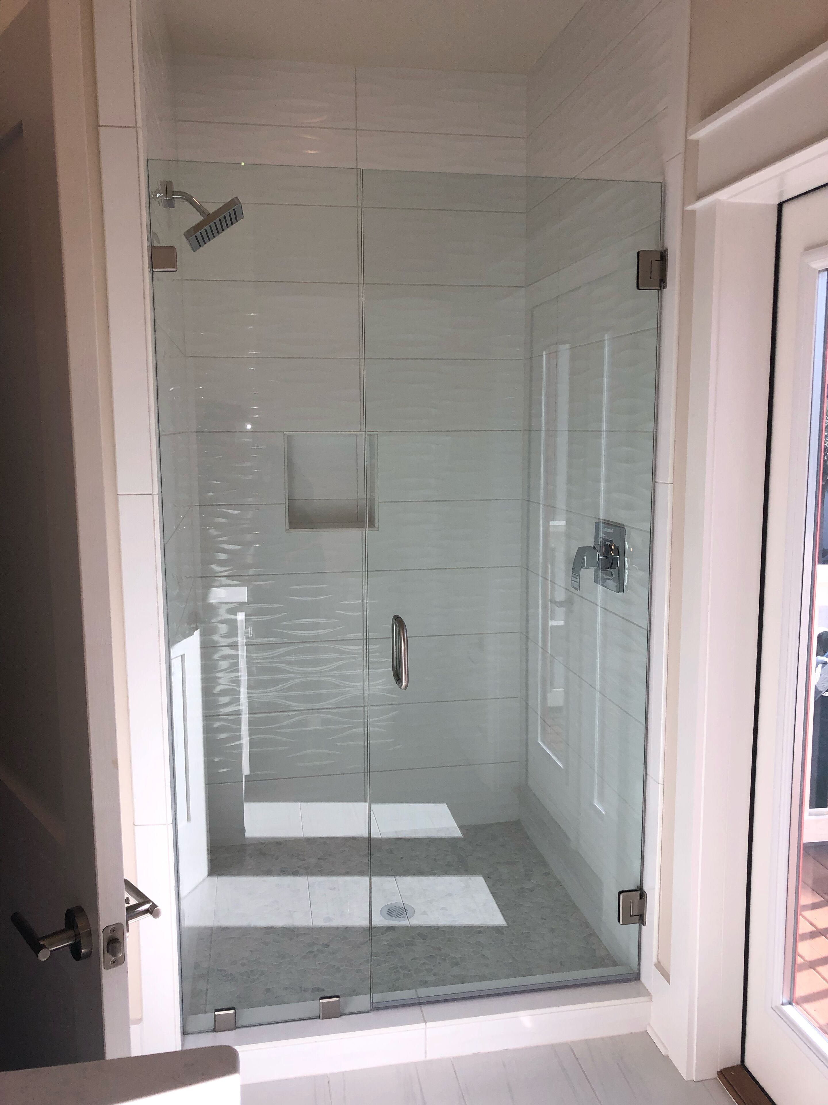 Une douche fermée par des parois en verre, dotée d’un pommeau de douche à main, d’un panneau de commande et d’un petit miroir carré.
