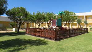 Un parque infantil con tobogán y estructuras para escalar, rodeado por una valla, con árboles y un edificio al fondo.