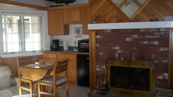 Une cuisine avec des meubles de rangement en bois, une cheminée en briques, une table à manger avec des chaises et un téléviseur fixé au-dessus des meubles.