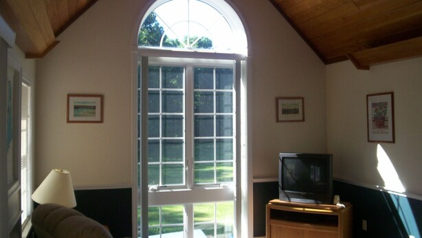 A room with a large arched window, a television on a wooden stand, and a lamp on a table. - Cool off in the Sunday River! Kayak to the Androscoggin. Spacious  Bethel home (Bethel)