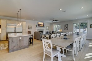 A spacious kitchen with a central island, a dining area with a long table and white chairs, a living room with a sofa and TV, and large windows with a view.