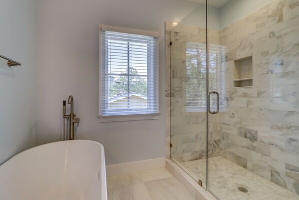 Un baño moderno con ducha envuelta en cristal, bañera y ventana con persianas.