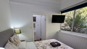 Un dormitorio con cama, televisión montada en la pared, ventana con vista a árboles y puerta que da a un baño.