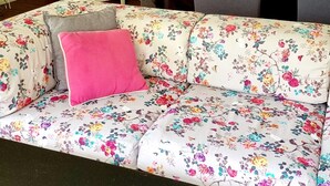 Una sala de estar con un sofá floral, una mesa de comedor con sillas, una planta en maceta y una ventana con persianas.