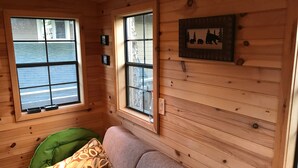 Ein gemütliches Blockhaus-Interieur mit Holzwänden, einem grünen Kissen auf einer beigen Couch und zwei Fenstern mit Jalousien.