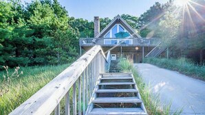 A wooden staircase leading to a house with a balcony, surrounded by greenery.