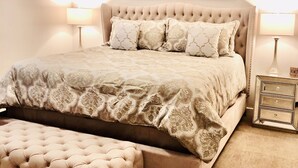 Un dormitorio con cabecera capitonada, una cama grande con ropa de cama estampada, un reposapiés a juego, dos lámparas de noche y un ventilador de techo.