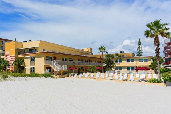 Ein Strandhotel mit mehreren Etagen, Balkonen und einer Strandlounge mit Liegestühlen und Sonnenschirmen.