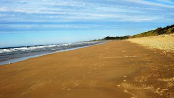 A wide sandy beach with gentle waves, a clear sky, and a distant shoreline with vegetation.