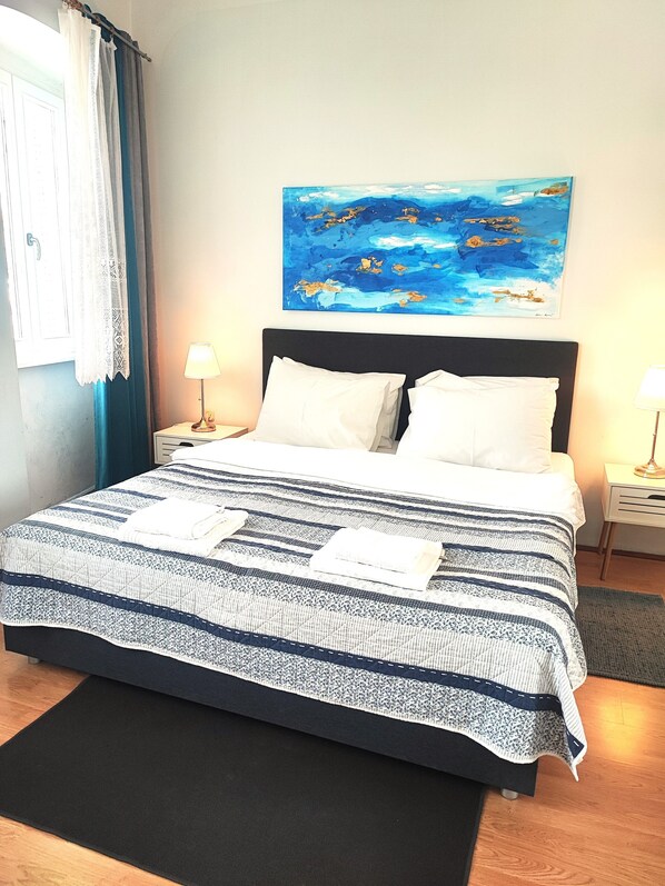 A neatly made bed with a striped bedspread, two white pillows, and two folded towels on the bed. There is a painting of a blue landscape on the wall above the bed.
